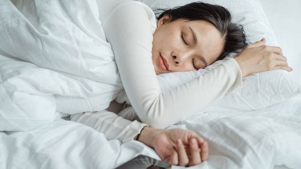 A woman in a white shirt sleeps in a bed with white ben linen.