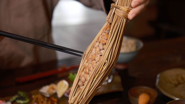 Natto in a straw holder, with chopsticks pointing towards it. 