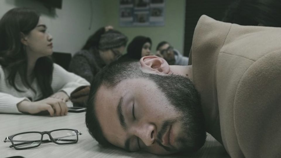 A person asleep with their on the desk with the other around are awake and chatting.
